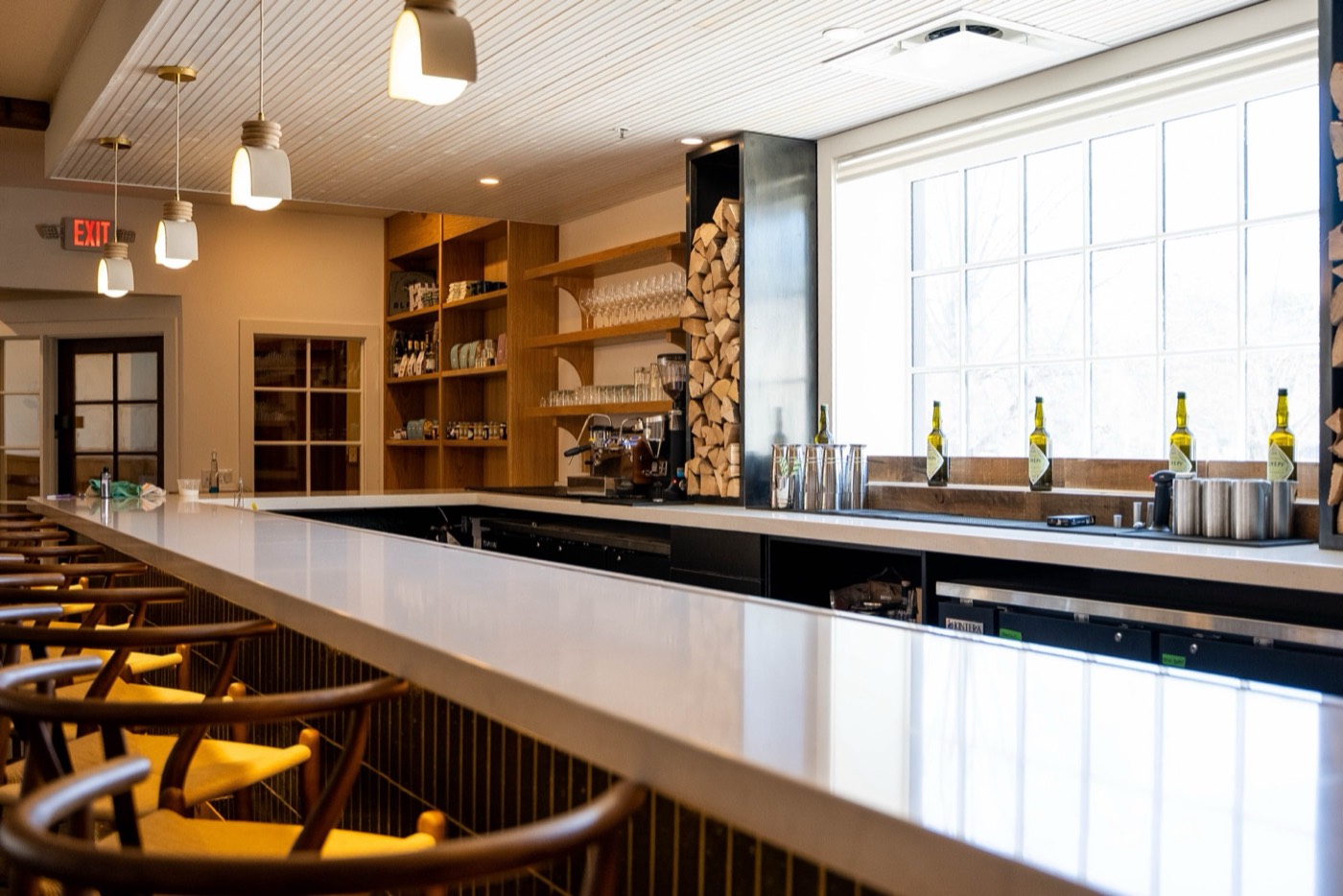 A finished bar build-out designed by Coffeehaus, wood shelving, stacked firewood, pendant lights, and a row of bottles ready for service